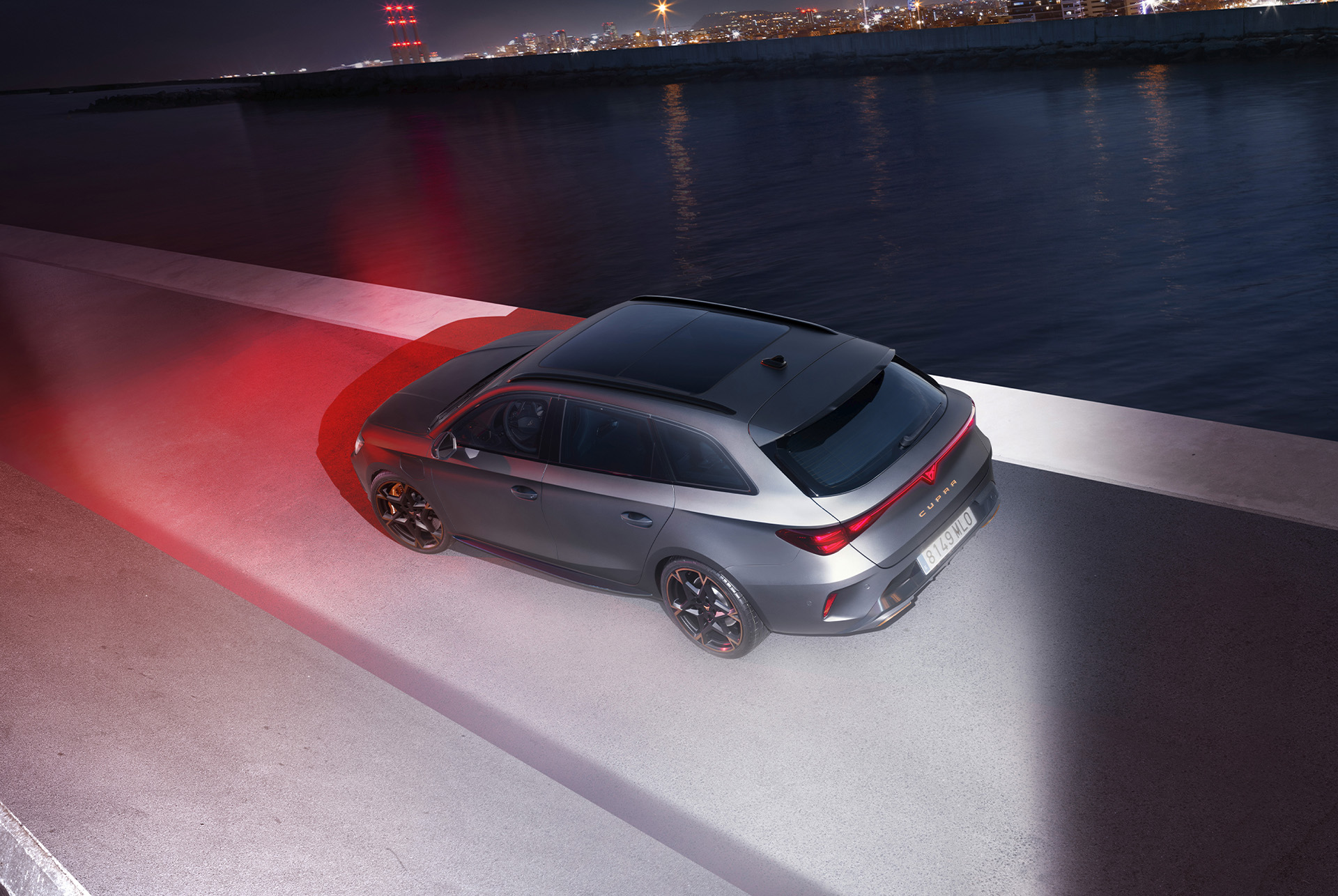 3/4 rear view of the CUPRA Leon SP in Magnetic Tech Matt Grey with 19-inch alloy wheels in Polar Copper, at night, in a urban seaside landscape.
