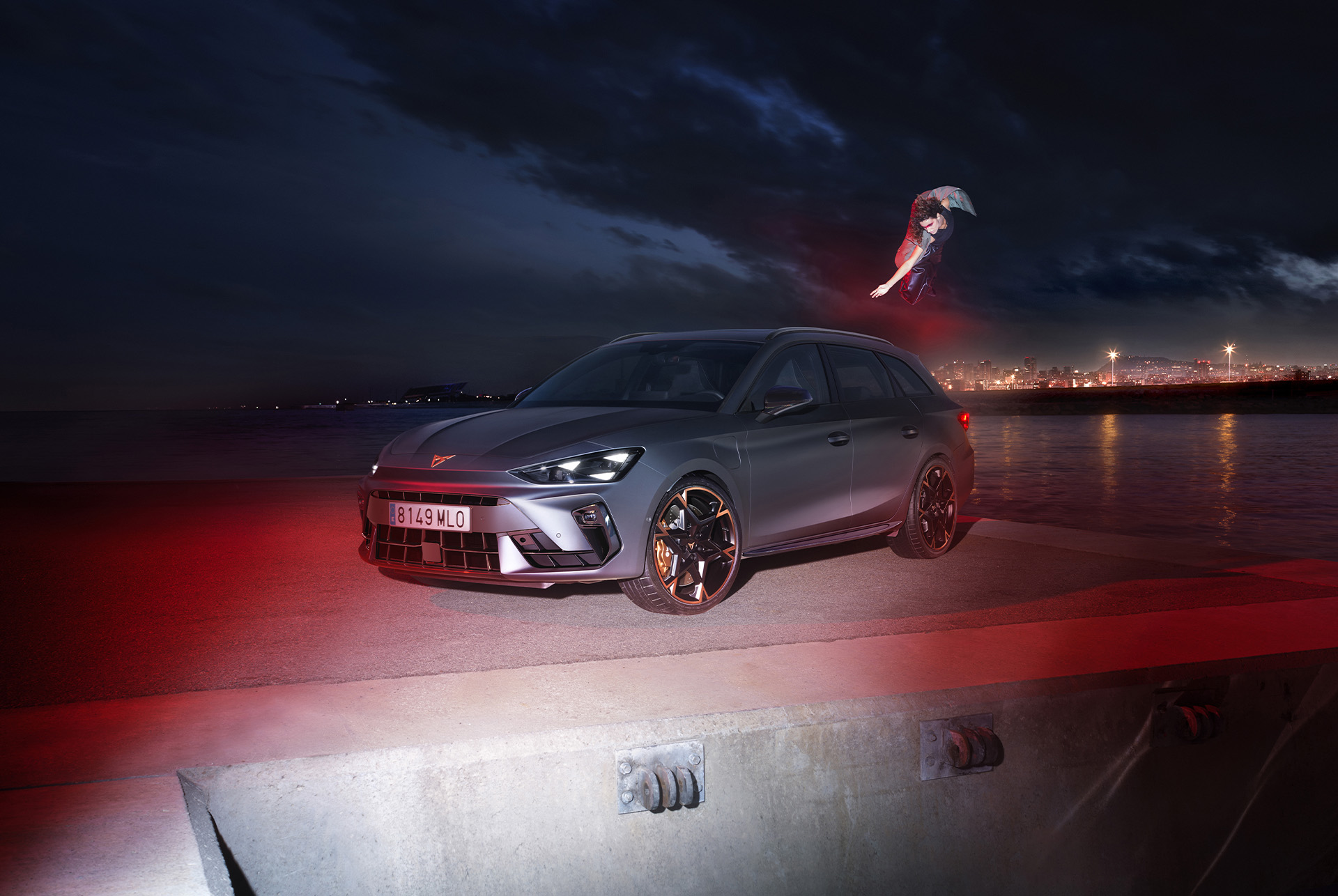 3/4 front view of the CUPRA Leon SP in Magnetic Tech Matt Grey with an urban seaside backdrop at night, showcasing 19-inch alloy wheels in Polar Copper. There's a person near the car.