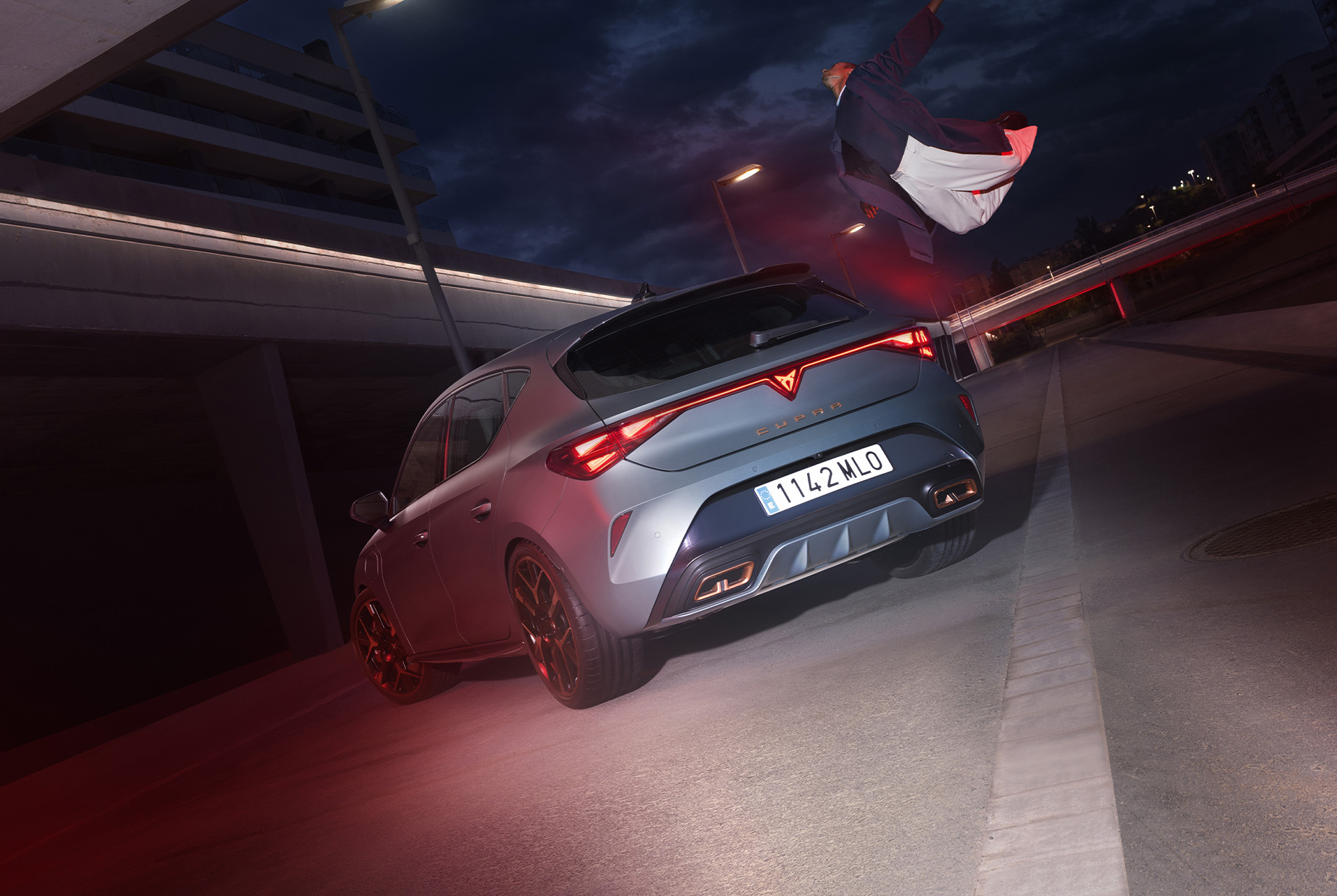 hero angle, 3/4 rear view of the CUPRA Leon PA 5D in Enceladus Grey Matt. The vehicle is parked in a urban setting at night, and there's a person next to it.