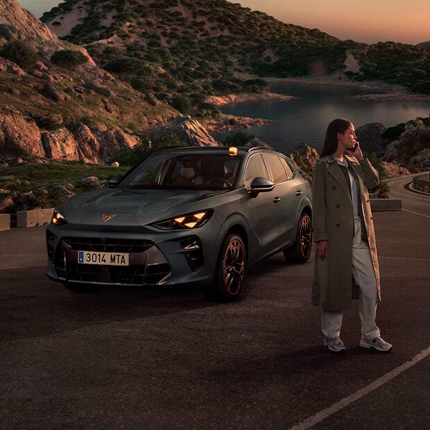 A CUPRA vehicle parked on a coastal road at sunset, with a person wearing a trench coat standing next to it, looking towards the sea.
