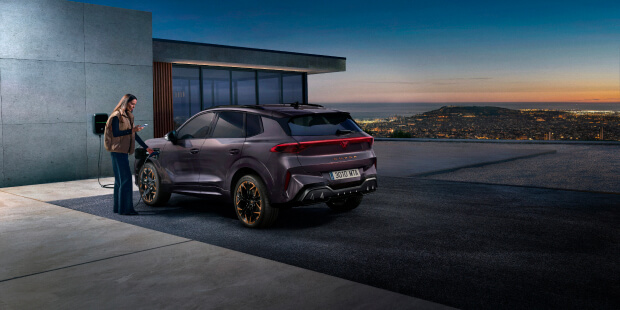  Woman interacting with the My CUPRA App next to a CUPRA Terramar hybrid SUV at a home charging station, highlighting smart connectivity for EV owners.